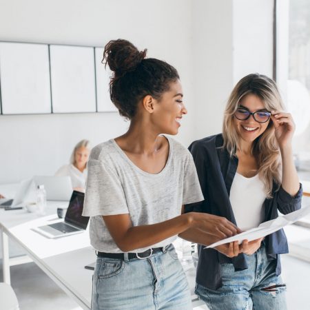 Young female executive explaines new strategy to blonde employee in glasses and smiling. Indoor portrait of multicultural collective working on project in office and using laptop.